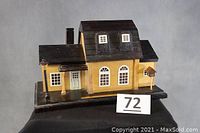 Wooden house model on display showing front and left side with yellow walls, black roof, white windows, chimney, porch, and birdhouse.