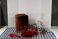 Set of six red and gold iced tea tumblers alongside white enamel coffee pot, red pottery baking dish, crocheted napkin holder, and basket on a blue and white checkered cloth.