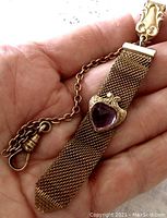 Hand holding antique gold-tone pocket watch chain with attached mesh fob featuring a heart-shaped glass amethyst stone.
