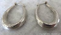 Pair of vintage sterling silver hoop earrings shown lying flat on cloth background, side views showing detailed engravings and oval hoop shape.