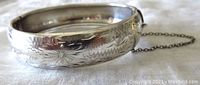 Close-up of bangle showing detailed floral engraving and polished silver surface with safety chain attached.
