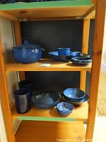 Wide view of the wooden shelving unit holding the lidded pot, the blue ceramic vases, bowls, and cups arranged on two shelves.