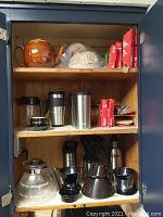 Cabinet shelf showing a brown ceramic teapot, several boxes of Melitta coffee filters in red and green packaging, stainless steel to-go mugs, a clear glass coffee pot, and multiple black plastic coffee funnels and drippers.