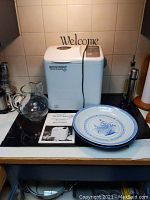 CounterCraft white automatic bread maker with digital controls, large blue and white ceramic plate, clear glass pitcher, and instruction manual on stovetop.