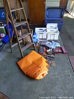 Overall lot layout showing wood ladder, orange tarp folded, several plastic bins, various hand tools and boxes of hardware on concrete floor.