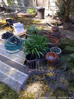 Photo showing assorted planter pots, garden hoses, wrought iron plant stands, and some greenery in an outdoor patio area.