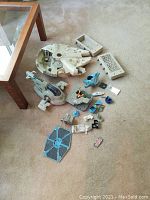 Wide view of disassembled and incomplete Star Wars toy parts on carpet including large Millennium Falcon top piece, smaller parts in blue, grey, white, and orange, and a blue TIE Fighter wing piece.