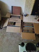 Three cardboard boxes filled with 78 RPM records visible packed inside, placed near a white brick fireplace and coffee table.