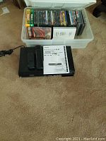 Assorted DVDs including unopened Harry Potter in a plastic storage bin with Toshiba DVD player and documentation on carpet.