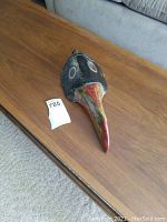 Wooden mask with long bird-like beak and circular eye highlights seen from top and side angles on wooden surface.