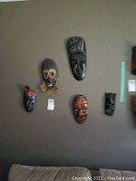Group photo of five hand carved wooden masks hung on a wall for display, varying sizes and styles mostly in dark and natural wood tones with some painted details and straw decoration on one mask.