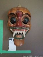 Front view of the wooden carved mask with large black bulging eyes and wide mouth with prominent white fangs.