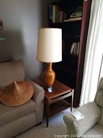 The full lamp shown in a living room setting beside a sofa and a light gray chair, showing the lamp with its fabric shade and ceramic body on a wooden side table.