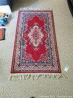 Full view of hand knotted area rug with red central medallion pattern and fringe tassels on the ends, laid out on beige carpet.