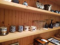 Shelf with various ceramic mugs, clear glasses, blue art glass cups, and a decorative tin.