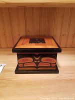 Front view of wooden box with painted Aboriginal design on the side and black trim framing the lid area.