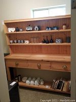 View of solid wood pantry shelving unit showing three upper shelves stocked with glassware, mugs, and decorative containers, three drawers below, and lower bottom shelf holding cups and books.