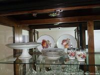 Glass cabinet shelf display showing Kuba Porzellan cake dish on pedestal, six fruit design plates stacked, and two Royal Worcester egg coddlers with metal lids