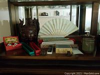 Full view of Asian decorative items displayed, including the wood carving, fans, containers, pottery dish, chopsticks in cases, and stamp arranged on a wooden shelf.