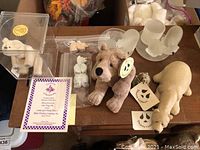 View of all eight polar bear figurines on a table including teddy bears with tags, frosted glass bears, carved stone bear, acrylic case with miniature bear and certificate
