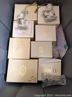 Gray tub with eight beige Lenox boxes with gold foil logo and two porcelain Lenox bear ornaments out of box resting atop boxes. Each box is beige with gold lettering 'Lenox' and gold emblem. Ornaments are white porcelain bears, one holding a present and wearing green scarf on sled, the other bear also on sled.