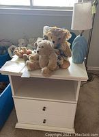 White nightstand with two drawers and open shelf holding teddy bears and blue ceramic table lamp.