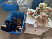 Photo showing numerous teddy bears in a blue plastic bin and on a small white table.