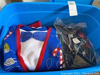 Blue tub containing a tightly folded woven knit Christmas sweater with a bowtie pattern and other holiday motifs, packaged with other items in a plastic bag.