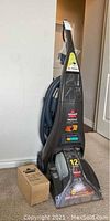 Full view of the Bissell ProHeat carpet cleaner standing upright with hose coiled behind and a small accessory box placed next to it.