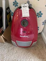 Red Simplicity Jill vacuum cleaner with visible brand logo and circular hose connection area, placed on carpet.