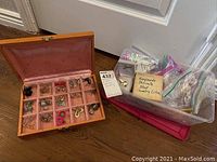 Overall view of two boxes: a pink velvet-lined wooden box with compartments containing various costume jewelry and a plastic bin with labeled baggies.