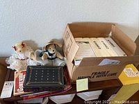Photo showing 2 collectible bears (one in white lace dress, one in blue outfit with a hat) alongside a box filled with magazines.