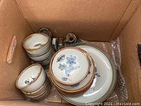 Box containing pottery set including plates, bowls, and salt and pepper shakers showing hand-painted blue bird and floral designs.