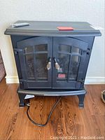 Front view of the black electric space heater designed like a stove with glass front doors and power cord on wooden floor.