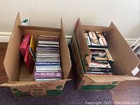 Two open cardboard boxes filled with various music CDs and DVDs placed on carpeted floor.