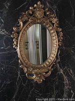 Front view of antique oval mirror with ornate gold plaster frame hung on a black marble wall showing floral and scroll details.