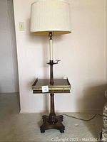Full view of the standing lamp showing the wooden base with carved details, square side table with brass edging, and beige pleated fabric shade on top.