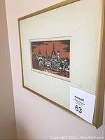 Framed woodcut print hanging on wall, shows red and black skyline image within gold metal frame and white mat