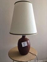 Full view of lamp on round table showing pottery base and light-colored tapered shade with gold trim.