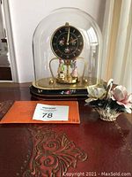 Front view of Kern German clock under glass dome with hand-painted base, porcelain figurines on the gold base plate, small porcelain flower basket to the right.
