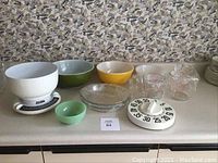 Full view of baking lot items on countertop including mixing bowls, glass measuring cups, pie plates, white kitchen scale, and mechanical timer.