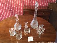 Overview photo showing two crystal decanters with stoppers, various small glasses, a small vase, and bowls on a wooden table.