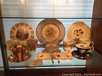 Display of multiple tea cups and collector plates with floral designs and gold accents arranged on shelf.