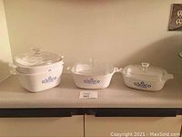Three Corning Ware casserole dishes with blue cornflower designs and glass lids arranged side by side on a counter showing different sizes.