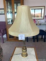 Full view of vintage table lamp with beige fabric shade and ornate brass base on marble-top table.