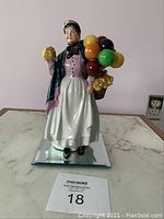 Front view of ceramic figurine of a woman holding balloons and a yellow object standing on a mirrored base