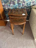 Photo showing three solid wood nesting tables stacked together with leather tops and gold embossing detail, positioned beside a sofa and floral wallpaper.