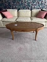 Full view of the oval coffee table placed on carpeted floor in front of a cream-colored sofa with pink pillows. Shows overall shape, glass top, and wood finish.