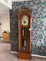 Front view of full grandfather clock against wallpaper, shows overall shape, height, finish