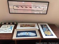 Framed cat chasing mouse print on wall and four art pieces on table below: ceramic tile, two signed prints, and feather bird print